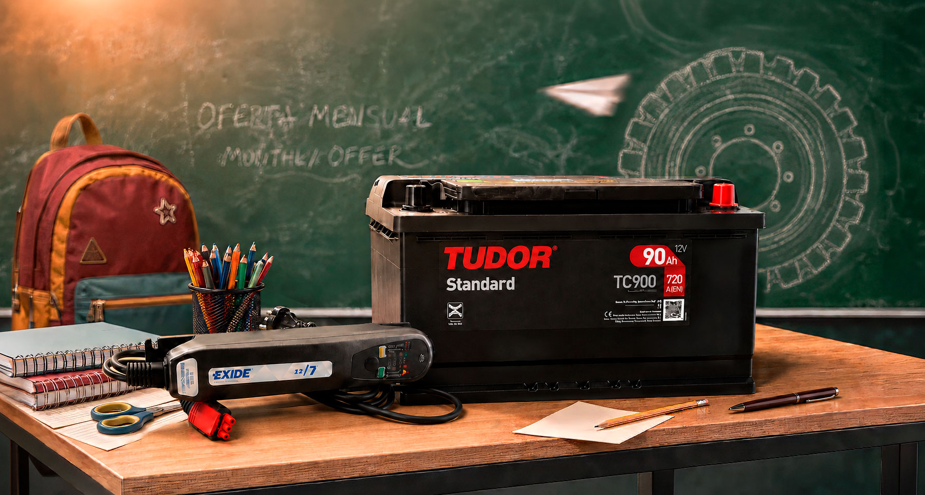 Batería TUDOR de 12 V y 90 Ah junto a un cargador EXIDE, sobre una mesa escolar frente a una pizarra con el mensaje «Oferta mensual.